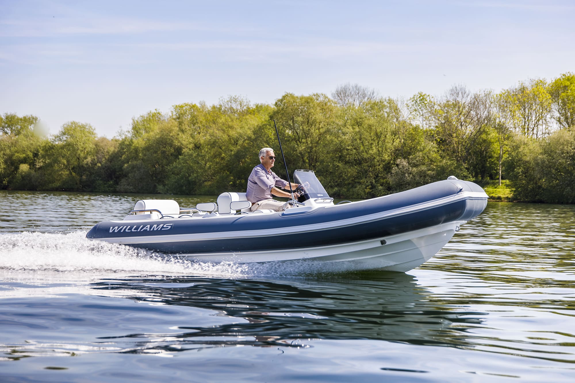 Man drives small inflatable boat on calm river with trees and blue sky background.
